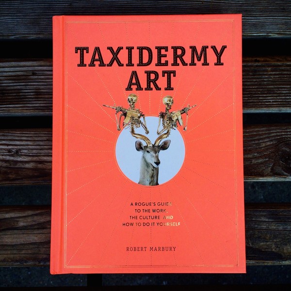Red book cover with gold foil details; photo in the center of a taxidermied deer with skeletons holding guns instead of antlers.