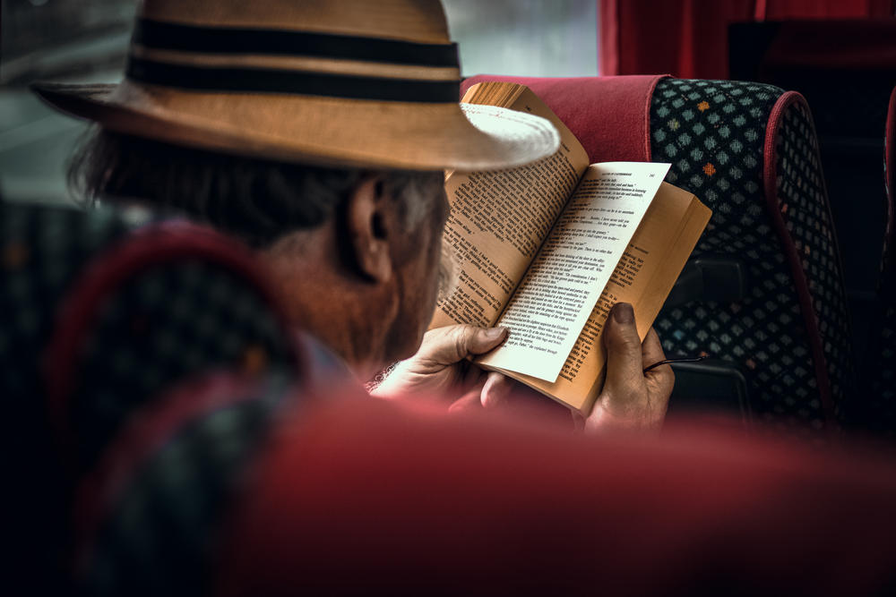 An over-the-shoulder photo of an older Black man reading a paperback book.