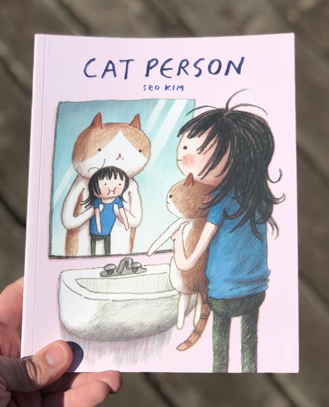A person holding a cat up to a mirror. In the reflection, the cat is pulling the person.