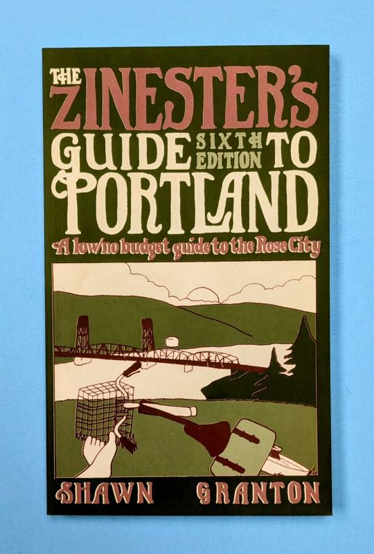 A green zine/small book with an illustration of a cyclist relaxing on a grassy hill, looking over the city of Portland and Hawthorne Bridge