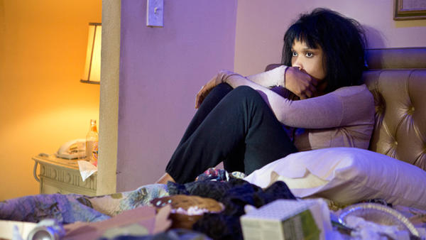 A movie still from Call Me Crazy showing a woman sitting on an unmade bed hugging her knees
