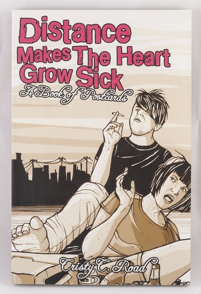 An emo looking couple sitting and smoking near the water with a pack of beer at their feet on a book cover