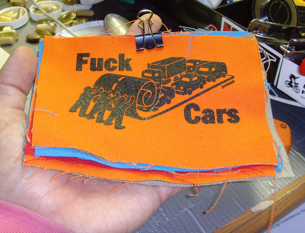 Three people rolling up the street like carpet while cars are on it. There is also the text “Fuck Cars” displayed. 
