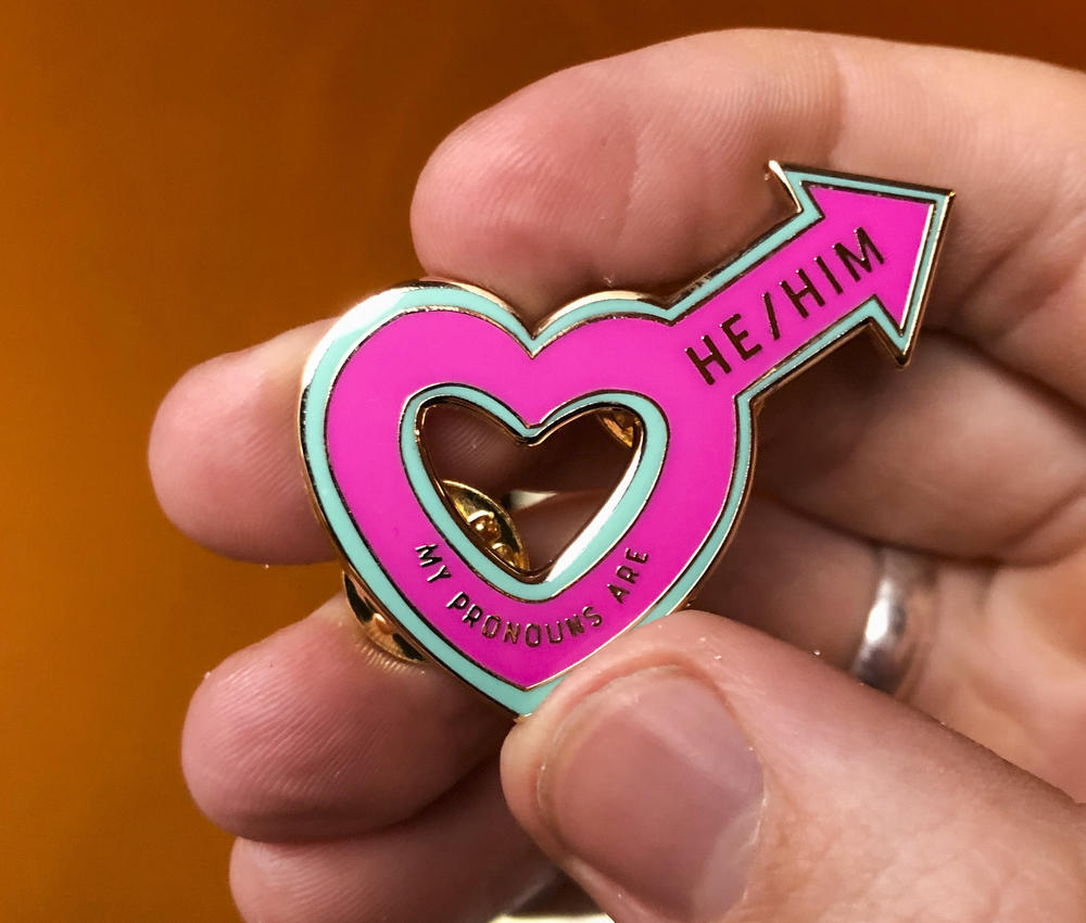An enamel pink male symbol in the shape of a heart with "he/him" written on it.