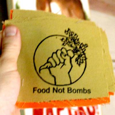 A hand holding a carrot with the text Food not Bombs printed on a canvas patch