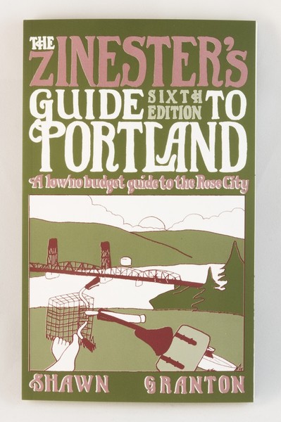 A green zine/small book with an illustration of a cyclist relaxing on a grassy hill, looking over the city of Portland and Hawthorne Bridge